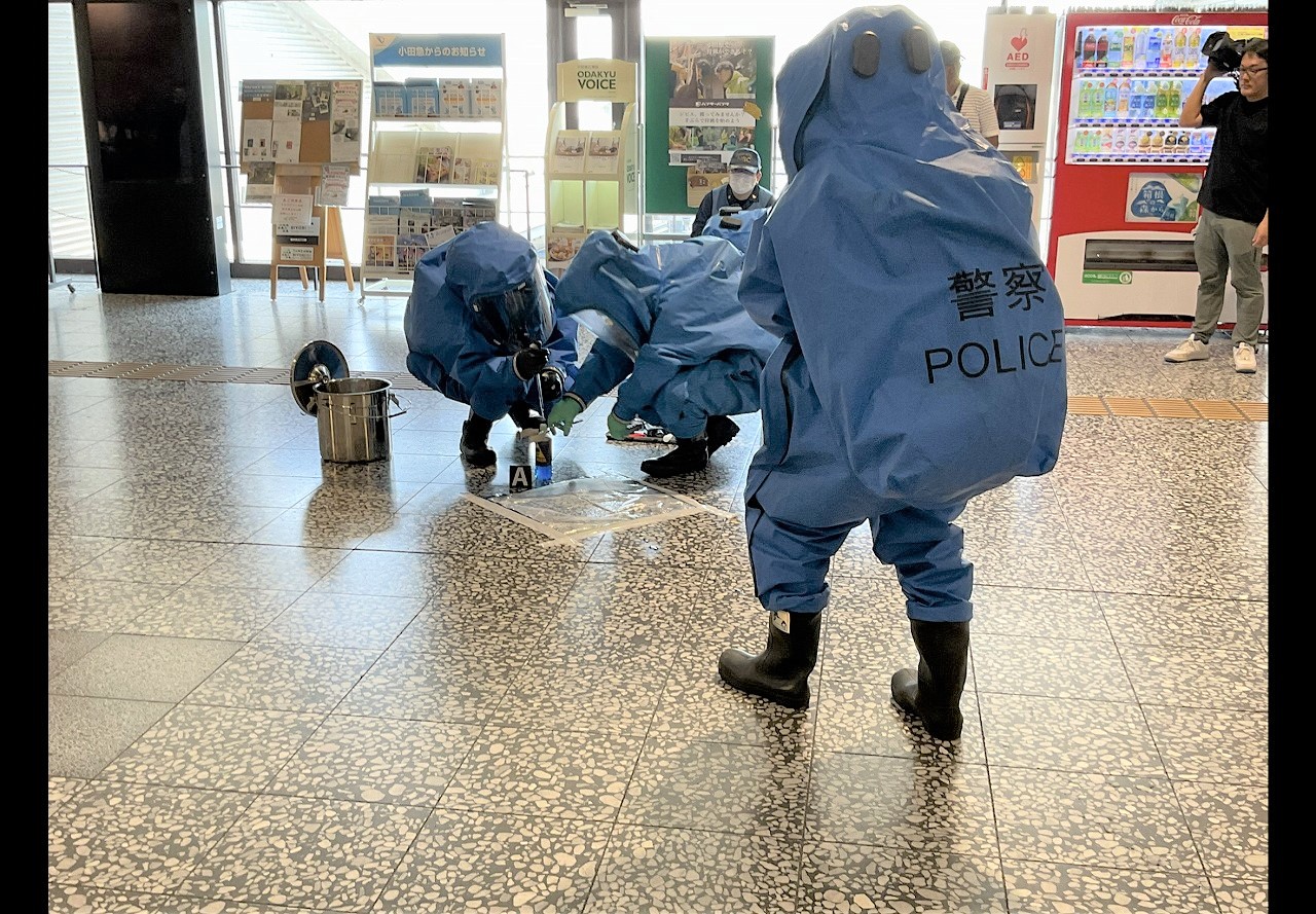 屋内駅コンコースのタイル床で、「警察 POLICE」と背中に書かれた青い化学防護服と全面マスクを装着した隊員三人が、黒い長靴を履き、銀色のバケツの横でビニールシート上の不審物らしきものにしゃがみこんで対応している緊迫した訓練の写真