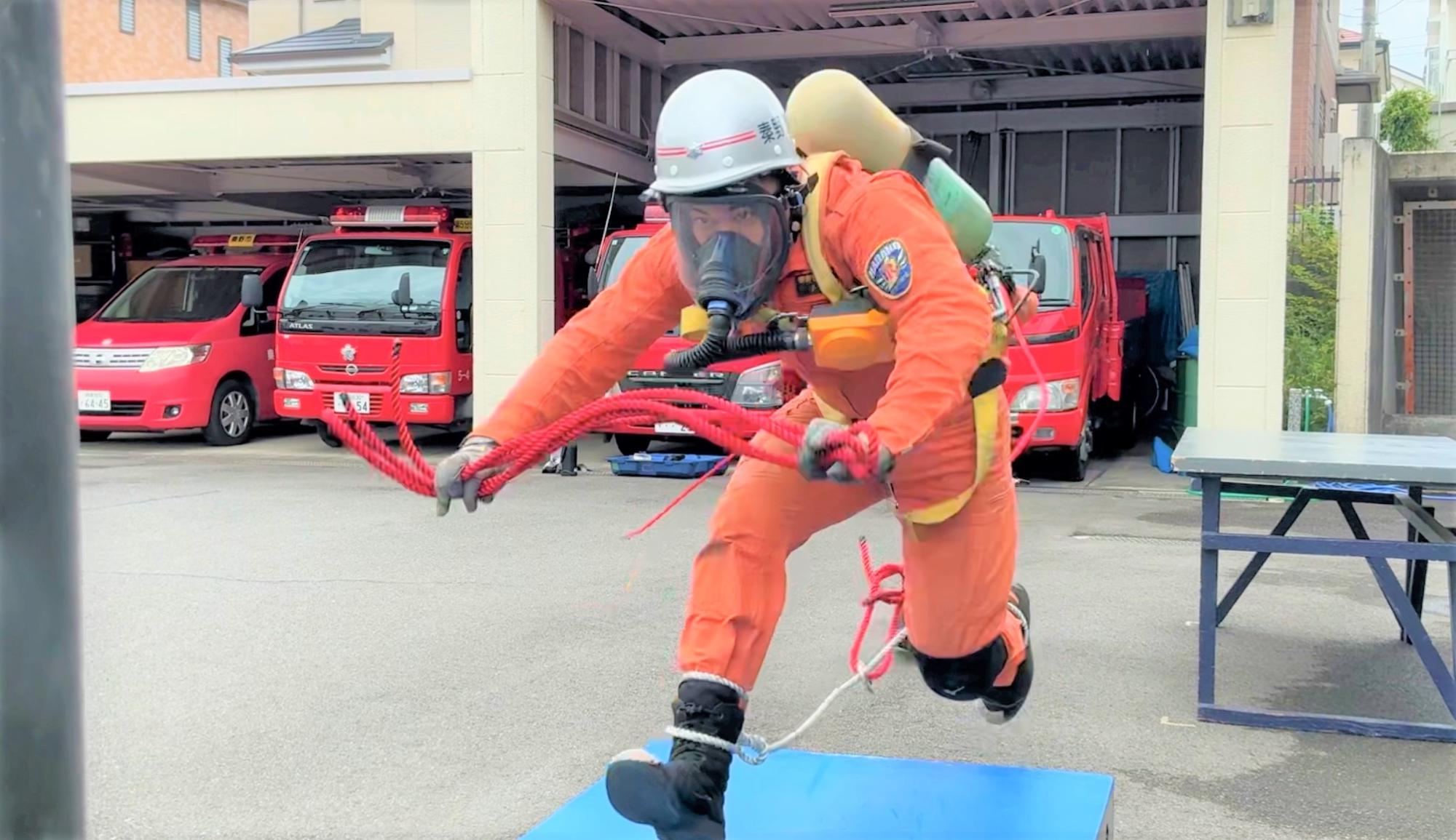 オレンジ色の救助服、白いヘルメット、酸素マスクを装着し、ボンベを背負った隊員が、赤いロープを手に持ち、走り出している様子の写真