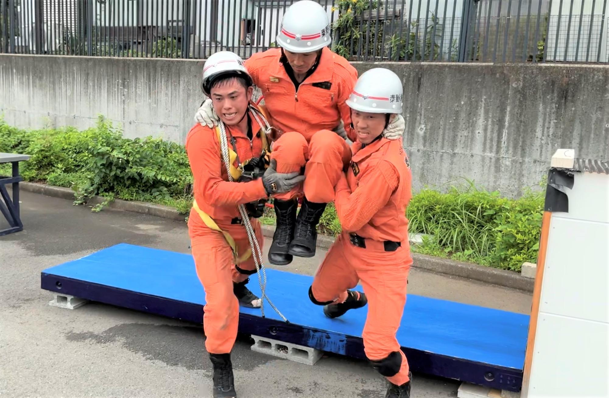 屋外の訓練場で、白いヘルメットとオレンジ色の救助服を着用した三人の隊員が、一人の隊員を二人で抱え上げるようにして搬送している様子の写真