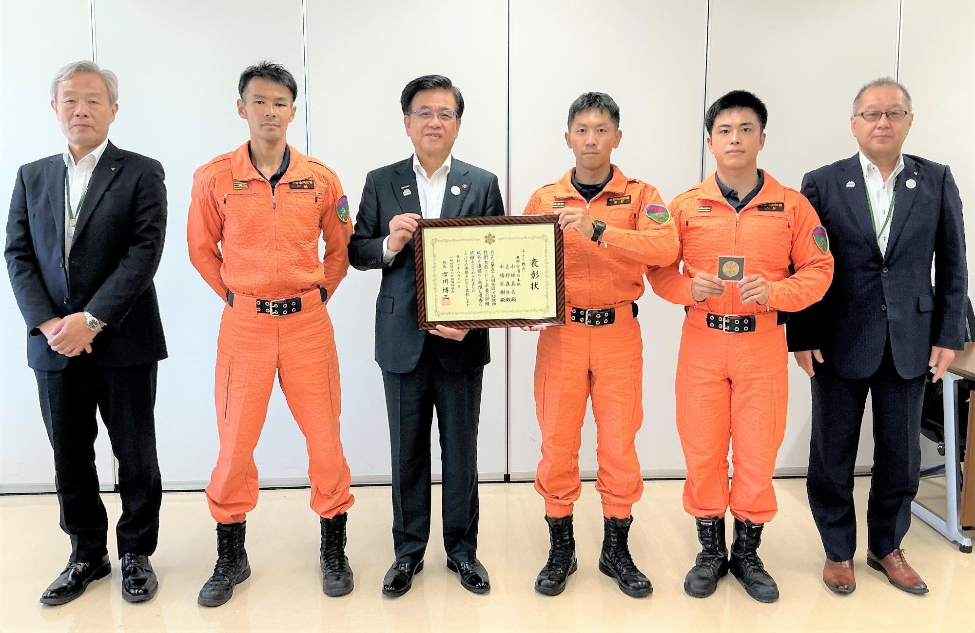 オレンジ色のレスキュー服を着た3人の男性とスーツ姿の3人の男性が横並びで並び、中央のスーツの男性とレスキュー服を着た男性が一緒に感謝状を持ち記念撮影している写真