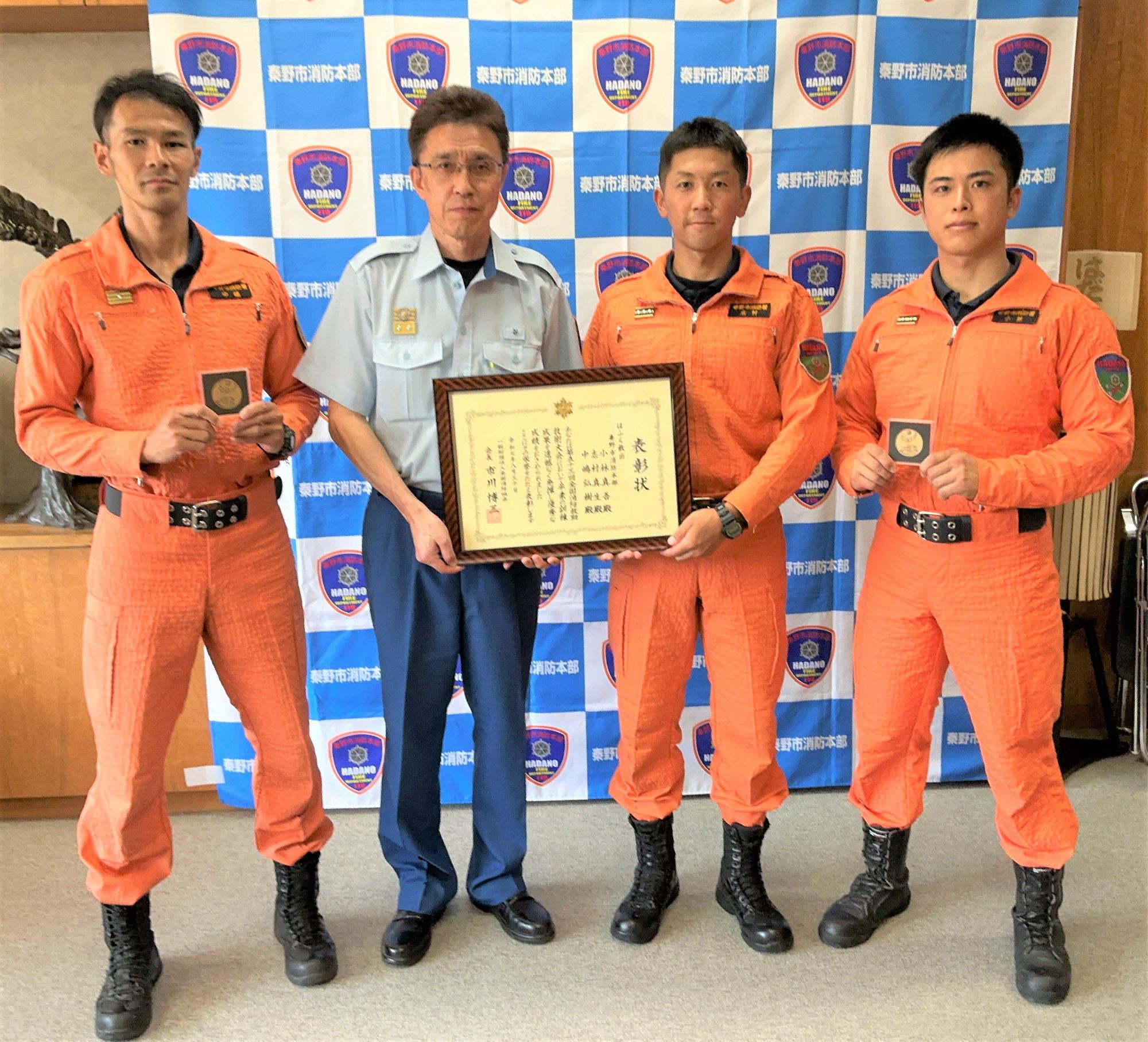 東野消防本部のバックボードの前で、オレンジ色のレスキュー服を着た3人の消防職員と制服姿の男性1人が、額縁に入った賞状を持っている写真