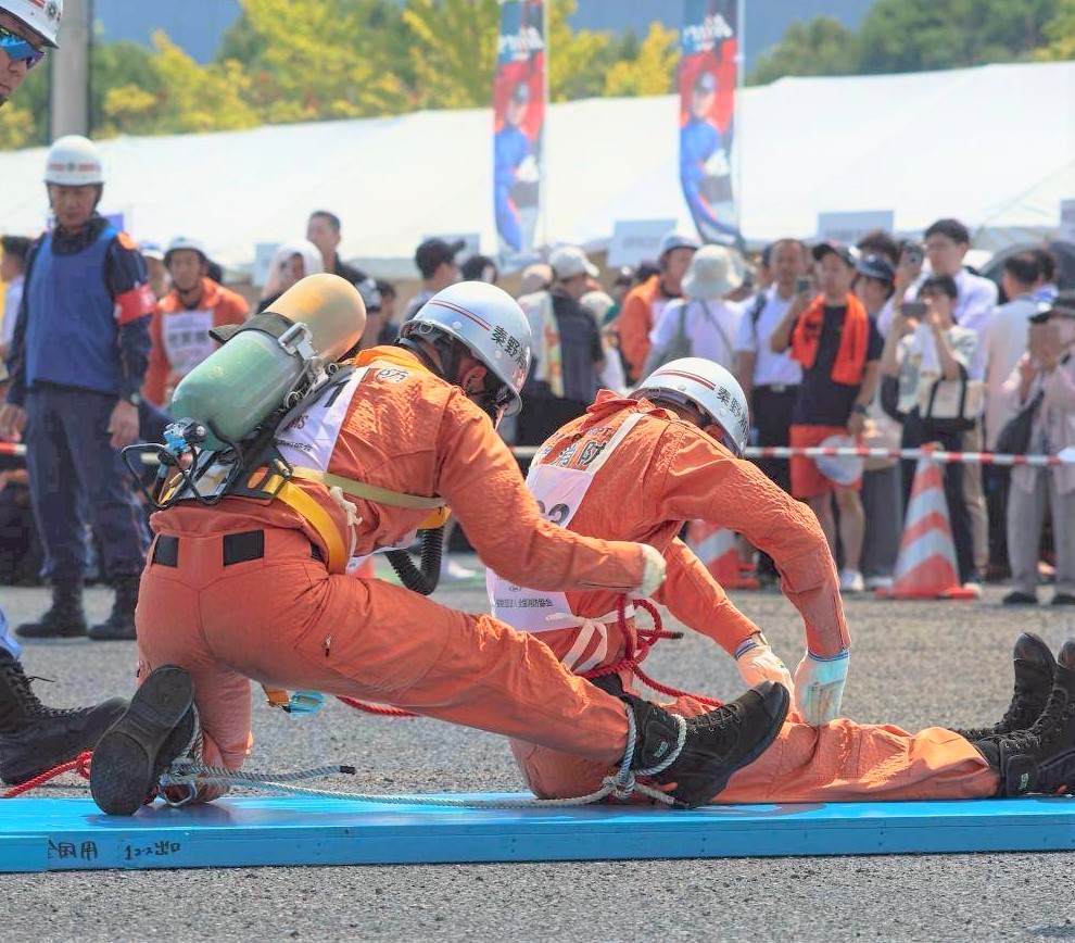 屋外で、レスキュー服を着てボンベを背負った隊員が、要救助役の隊員の腹部を後ろから抱きかかえて救助活動を行っている様子の写真