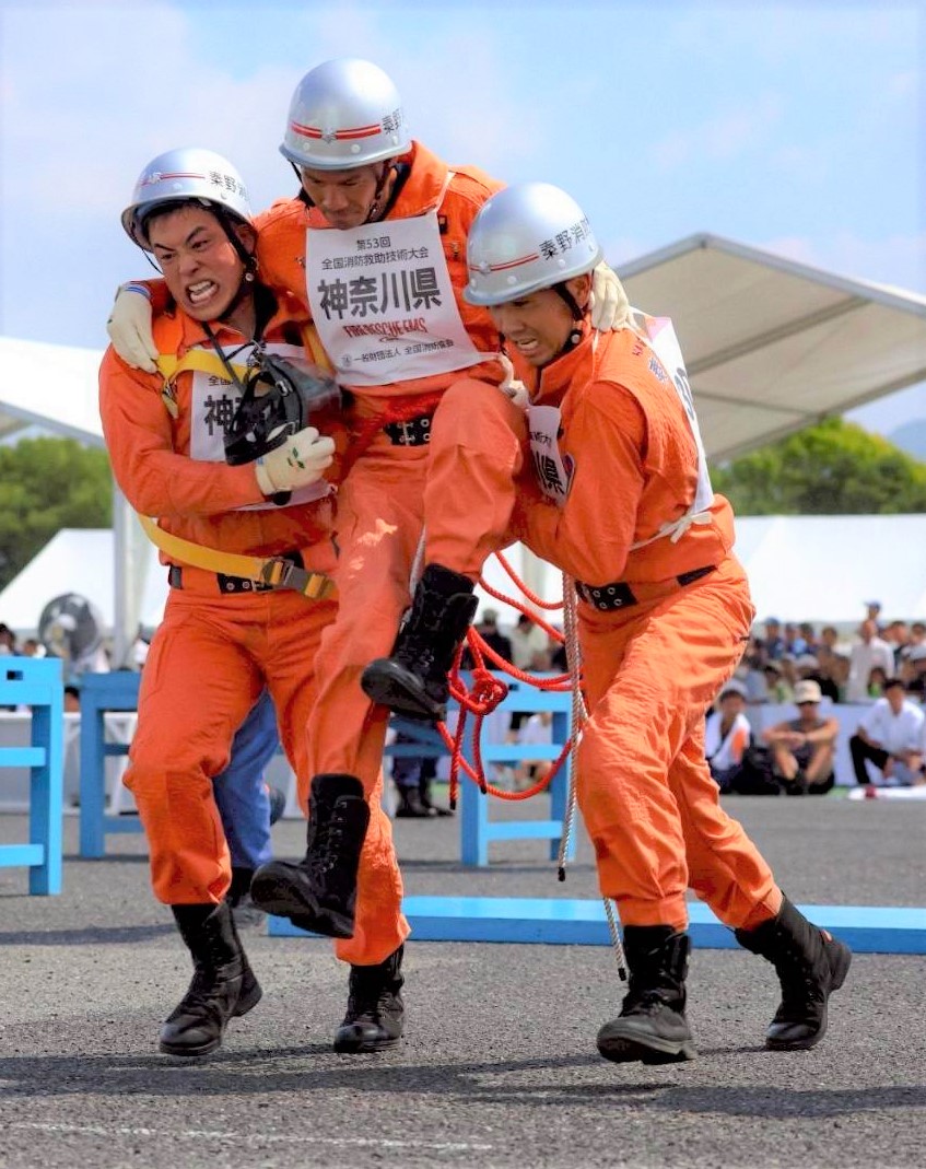 レスキュー服とヘルメットを着用した消防隊員3人が、訓練の最中に中央の負傷者役の隊員を左右の隊員が両側から抱え上げ救出している様子の写真