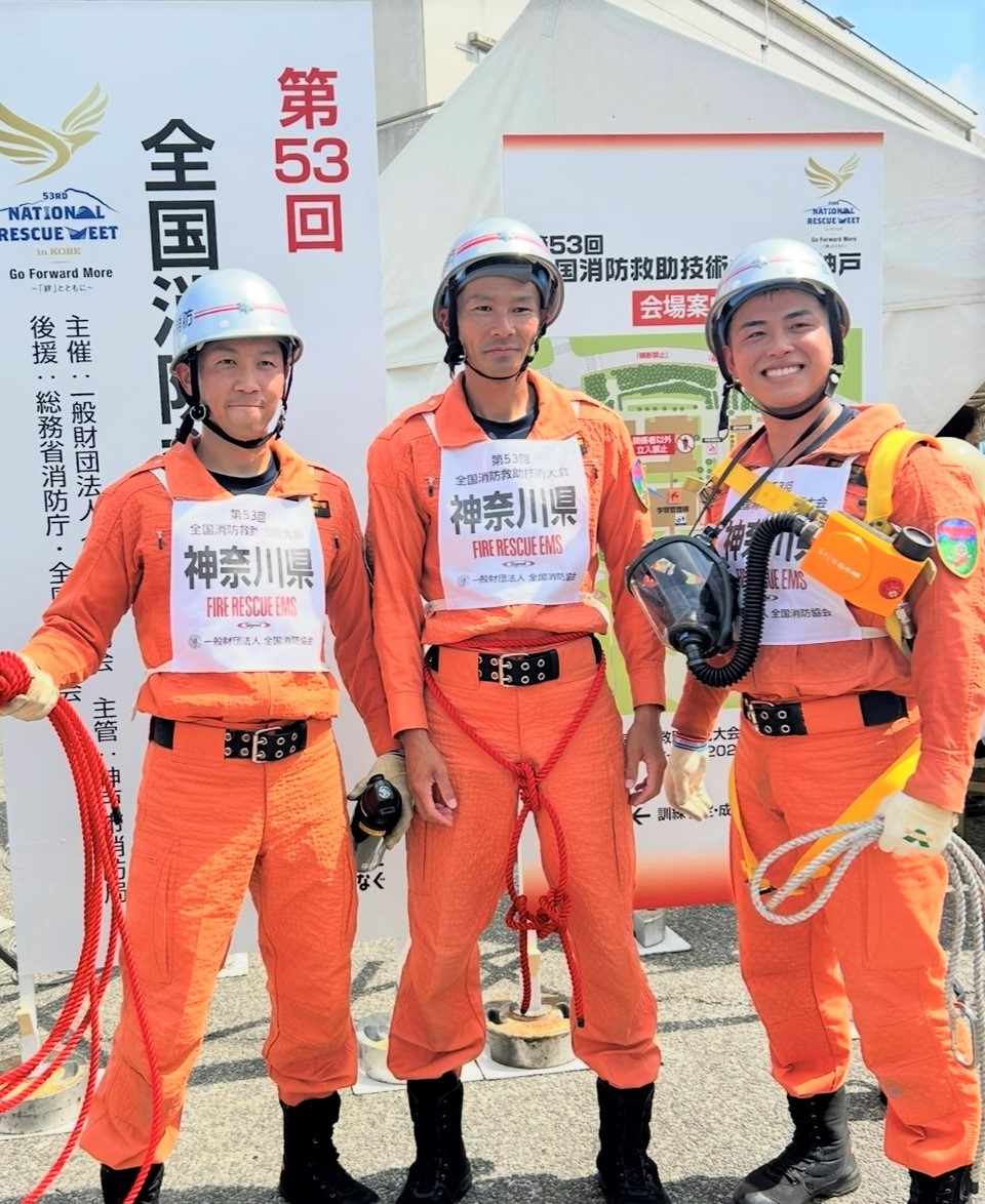 レスキュー服とヘルメットを着用した消防隊員3人が、背景にある「全国消防救助技術大会」の看板の前で、ロープや資機材を身につけて笑顔でポーズをとっている写真