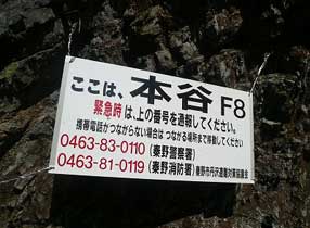 「ここは、本谷 F8」と記された現在地情報と、赤字で「緊急時は、上の番号を通報してください」の注意書き、そして秦野警察署と秦野消防署の電話番号が記載された緊急連絡用の標識の写真