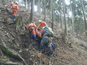 急峻で土砂がむき出しの斜面が広がる林の中で、オレンジ色の救助服を着た複数の隊員が、緑のバックパックを背負った山岳遭難救助隊と協力しながら、ストレッチャーに乗せた要救助者を搬送している山岳救助訓練の様子の写真