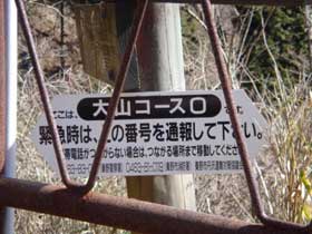 黒い丸の中に白抜きで「大山コース0」と書かれ、その下に「緊急時は、この番号を通報して下さい。」と続く注意書きと連絡先の電話番号が記載された登山道の案内兼緊急連絡先標識の写真