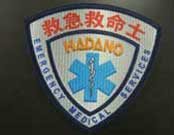 盾型で白地のワッペンで、青い星型生命のシンボルとアスクレピオスの杖が中央に描かれ、上部に救急救命士、中央にHADANO、下部にEMERGENCY MEDICAL SERVICESの文字が入った、救急救命士のエンブレム写真