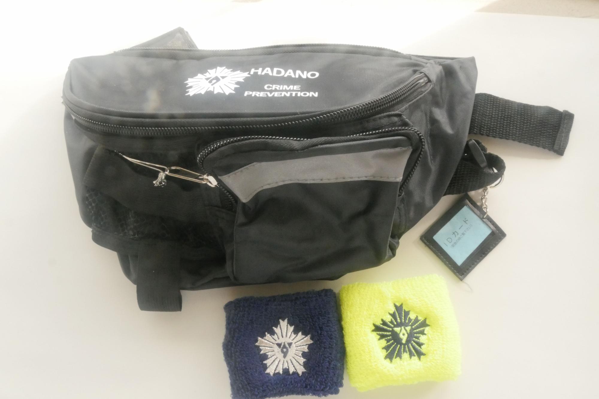 「HADANO CRIME PREVENTION」の文字とシンボルマークがプリントされた黒いウエストポーチ、同じシンボルマークが刺繍された紺色と蛍光黄色のリストバンド2つが置かれた隊員グッズの写真