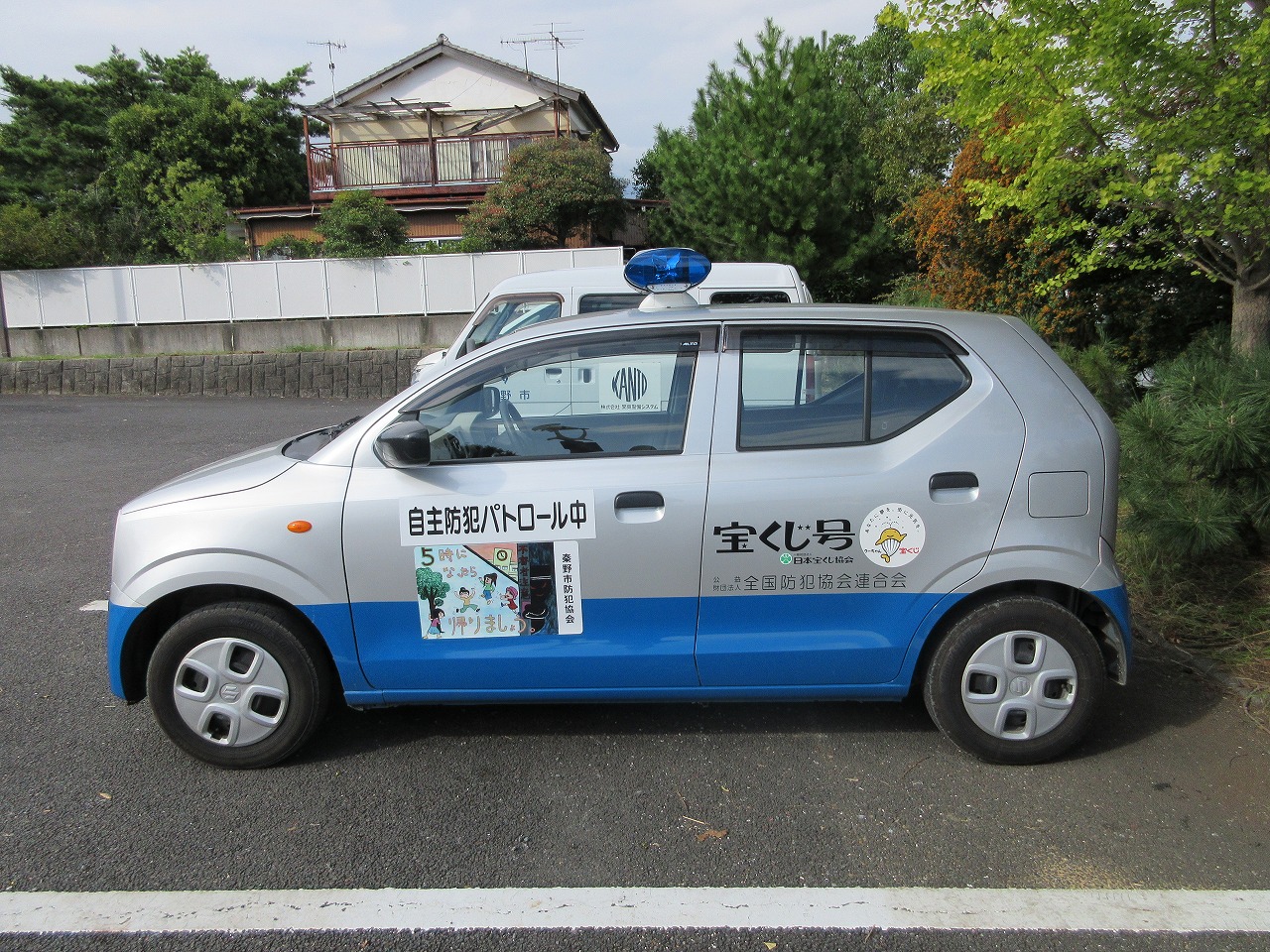 駐車場の舗装された地面に停められた、銀色と青色のツートンカラーで上部に青い回転灯を載せた青パトの写真