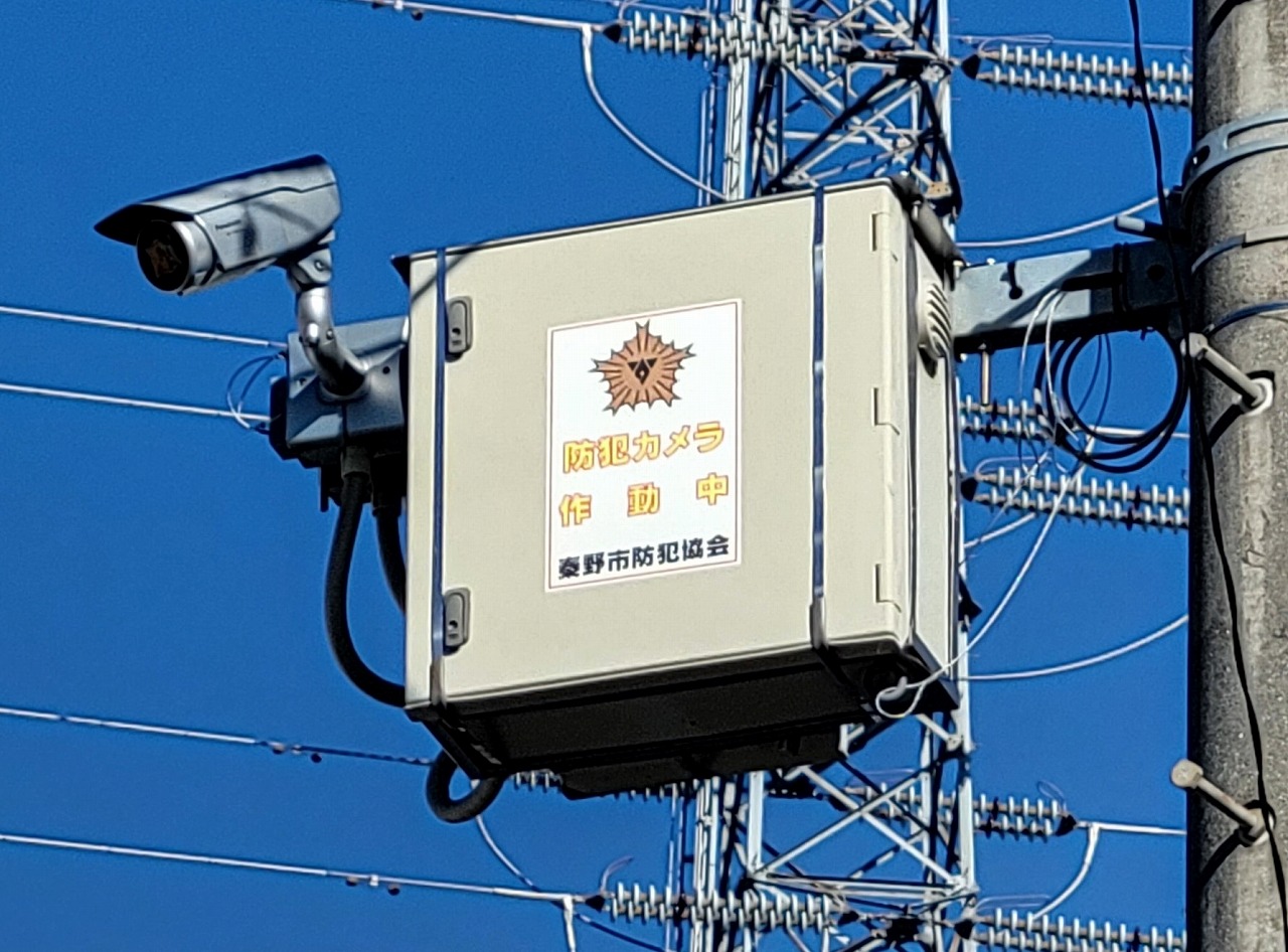 「防犯カメラ作動中 秦野市防犯協会」と書かれたグレーの制御ボックスと防犯カメラが電柱に取り付けられた写真