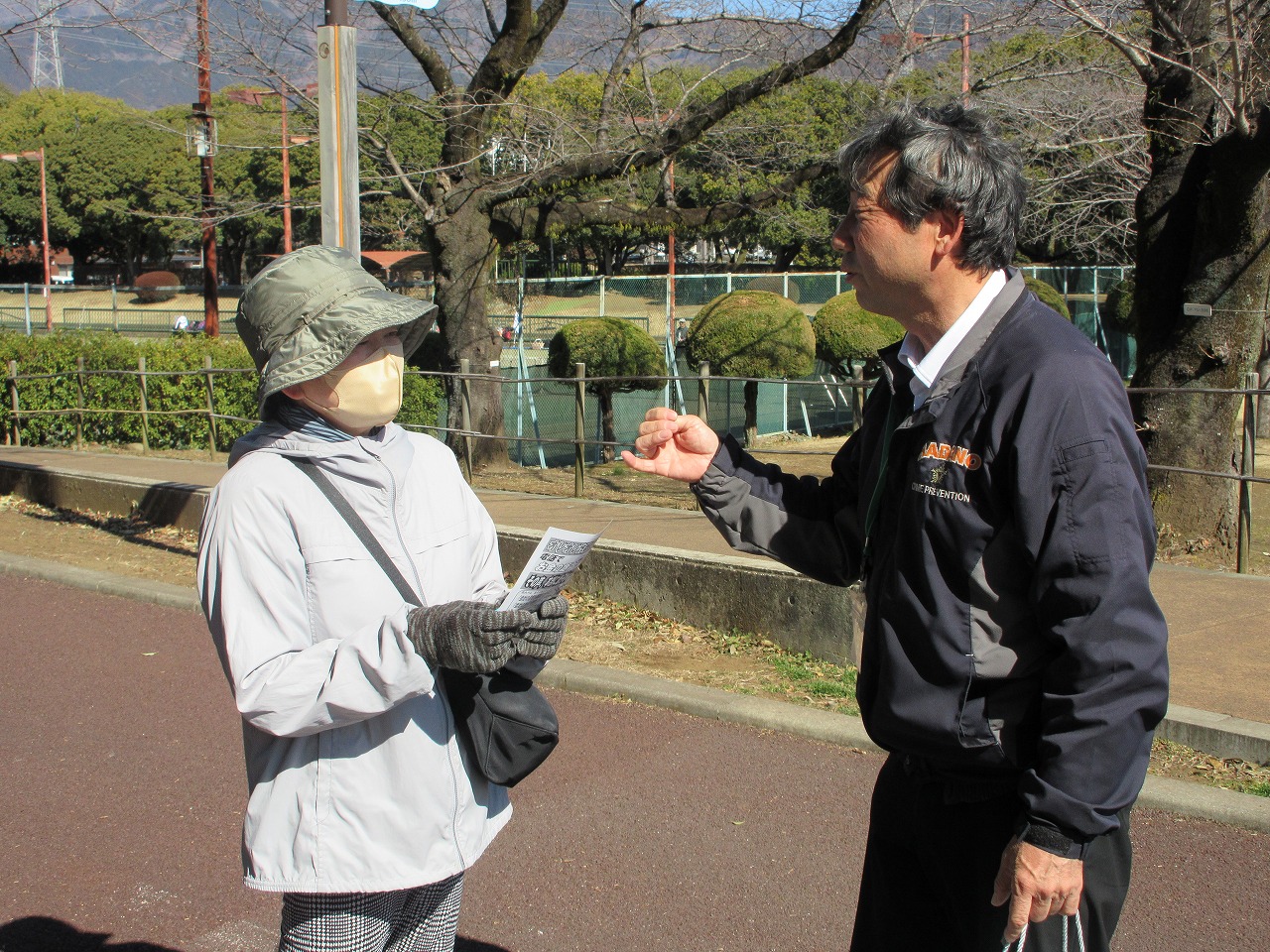 明るい日差しの当たる公園の舗道で、防犯パトロール用のジャンパーを着た男性が、帽子とマスクを着用した女性に対し、片手に持ったチラシのようなものを指さしながら話しかけている様子の写真