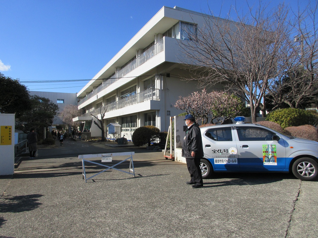 学校の正門前に、「自主防犯パトロール中」と書かれた銀と青のパトロールカーが停まり、制服姿の男性がその横に立って校舎を見ている様子の写真