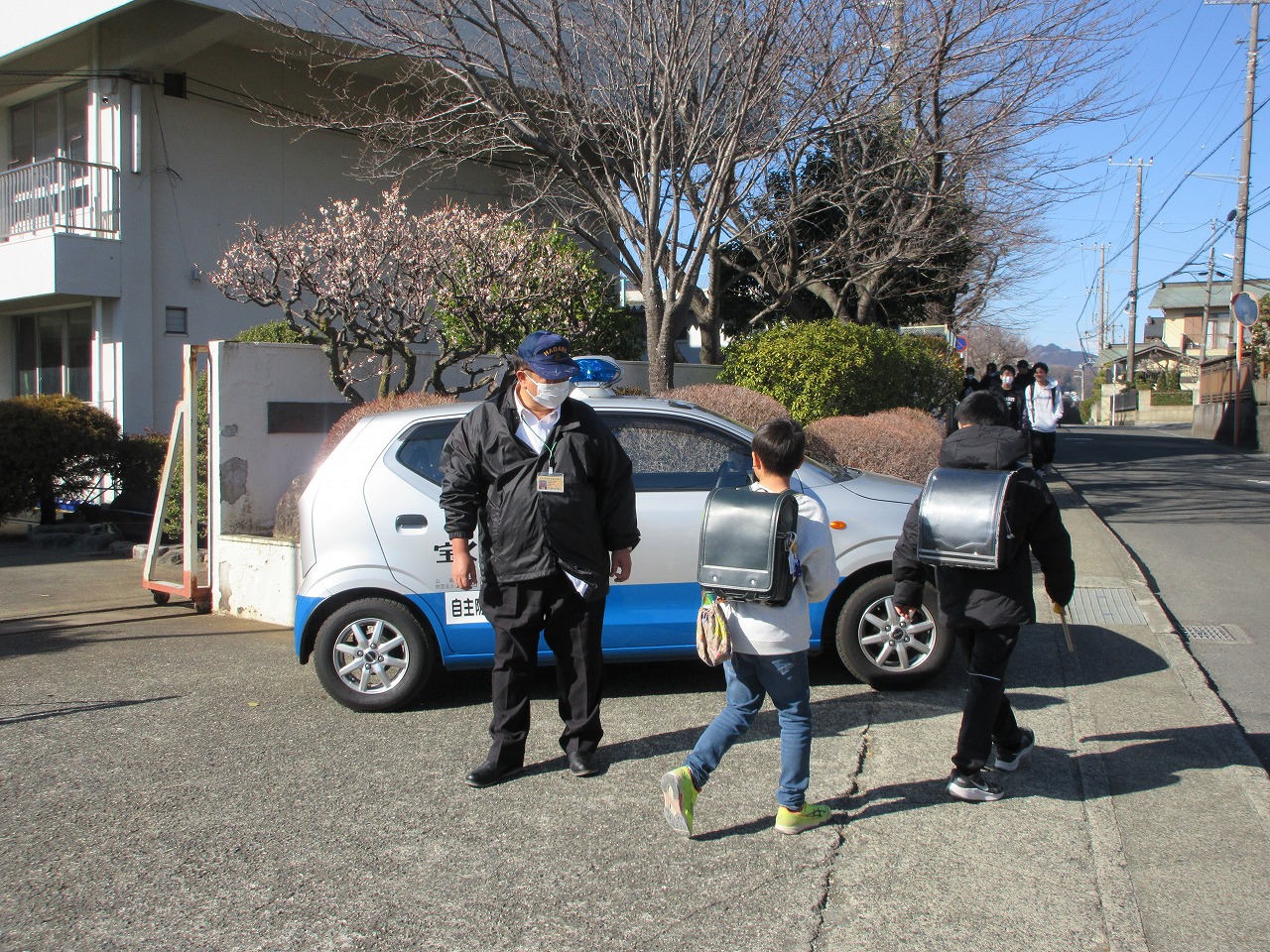 通学路の歩道に停めた自主防犯パトロールカーの横で、黒い上着とマスクを着用した制服姿の男性が黒いランドセルを背負った小学生たちを見守っている写真