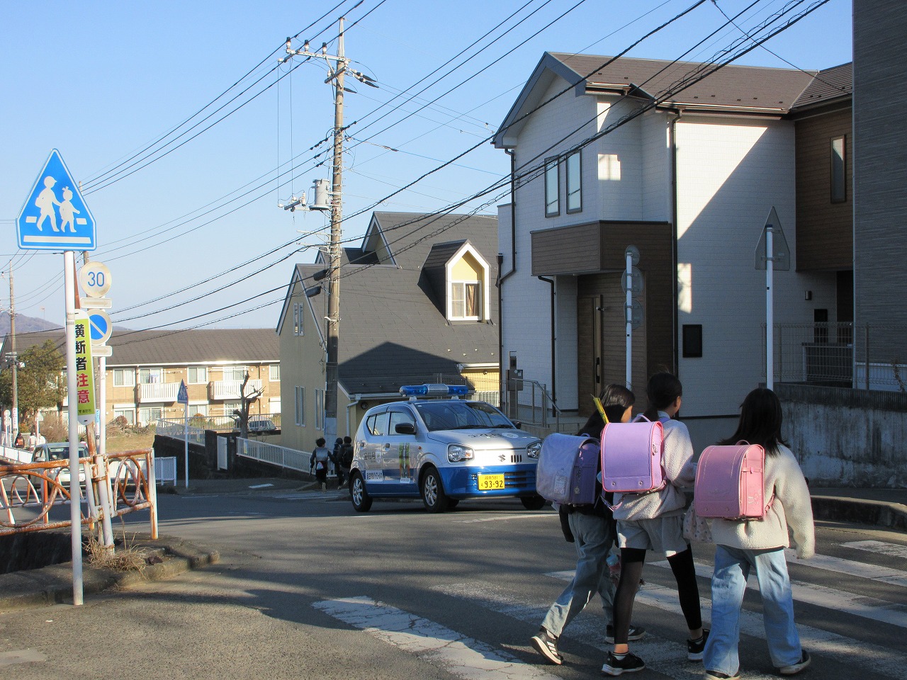 ランドセルを背負った小学校が住宅街の横断歩道を渡っており、背後に青い回転灯を載せたパトロールカーが見える写真