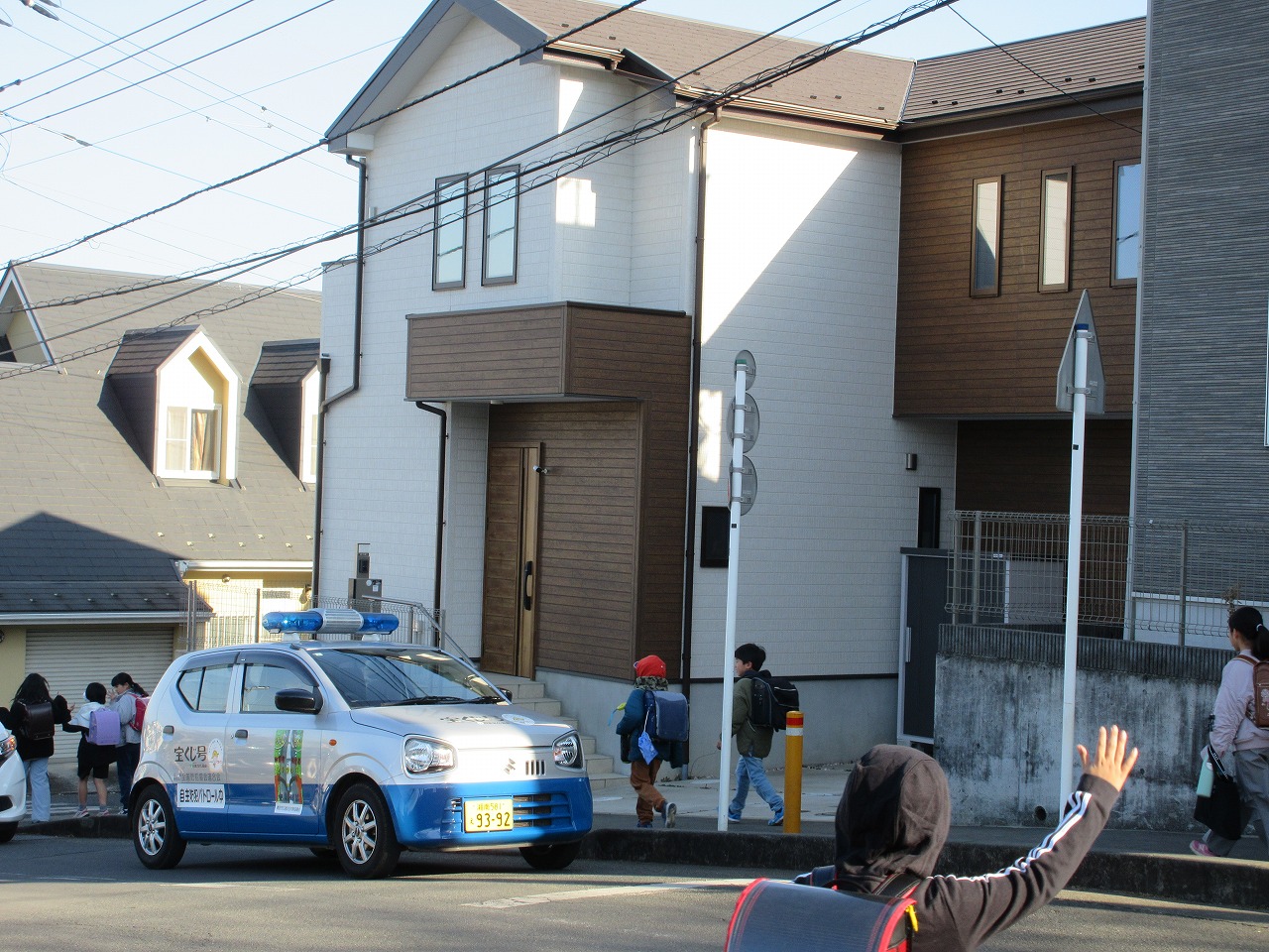 住宅街の道路に停車している青い回転灯を載せた自主防犯パトロールカーの横を、赤や緑のランドセルを背負った子どもたちが登下校している写真