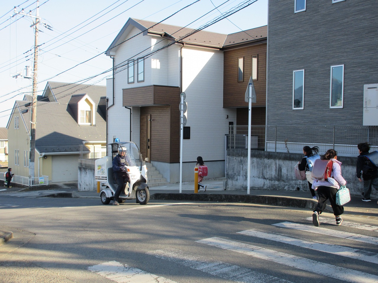 住宅街の交差点にある横断歩道付近で、青い回転灯を付けた白い三輪バイクに乗った人が、下校中のランドセルを背負った小学生たちを見守っている、防犯パトロールの様子の写真