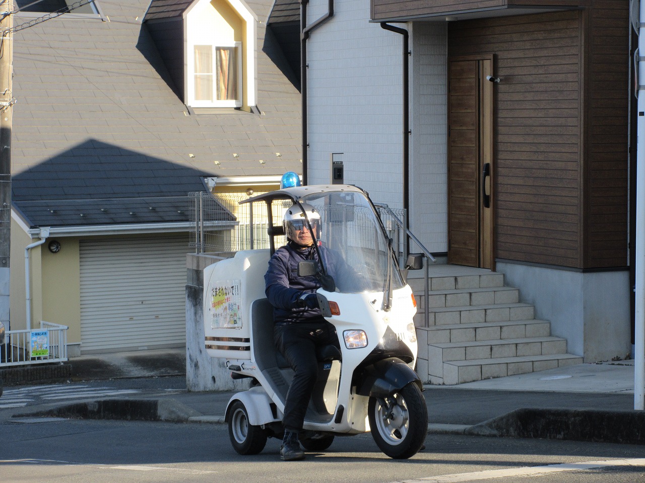 住宅街の道路上で、「見守り活動中」と書かれたボックスを装備し、青い回転灯を付けた白い屋根付きの三輪バイクに、ヘルメットを被った男性が乗っている写真