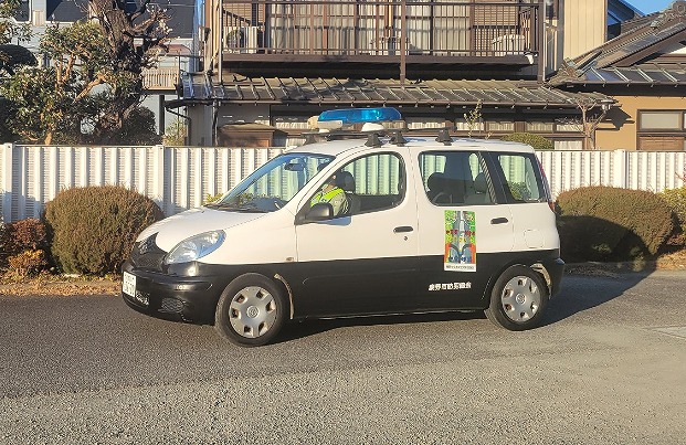 住宅街の道路に停車している青い回転灯を載せた、白と黒のツートンカラーのワゴン車で、側面に「藤野地区防犯協会」と書かれたステッカーが貼られている写真