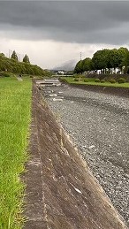 暗雲が垂れ込める空の下、石積みの護岸が整備された河川と緑の土手が広がる増水前の田園地帯の河川風景写真
