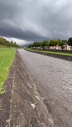 厚い雲が覆う曇天の下、石積み護岸の河川に濁った水が流れ、対岸に住宅地と緑が連なる大雨後の増水した川の様子を撮影した風景写真