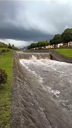 暗雲が迫る曇天の下、コンクリート護岸を濁流が激しく流れ落ち、堰堤から白い飛沫を上げる大雨後の増水した河川の危険な状況を捉えた防災記録写真