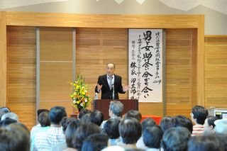 木目調の壁を背景に「男女共同参画」と書かれた白い掛け軸の前でスーツ姿の講師が演台で講演し、多数の参加者が着席して聞く男女共同参画推進セミナーの写真