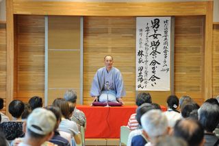 木目調の壁と「男女共同参画」の掛け軸を背景に、着物姿の講師が赤い毛氈の舞台で正座し、多数の参加者が見守る伝統文化体験を含む男女共同参画イベントの写真