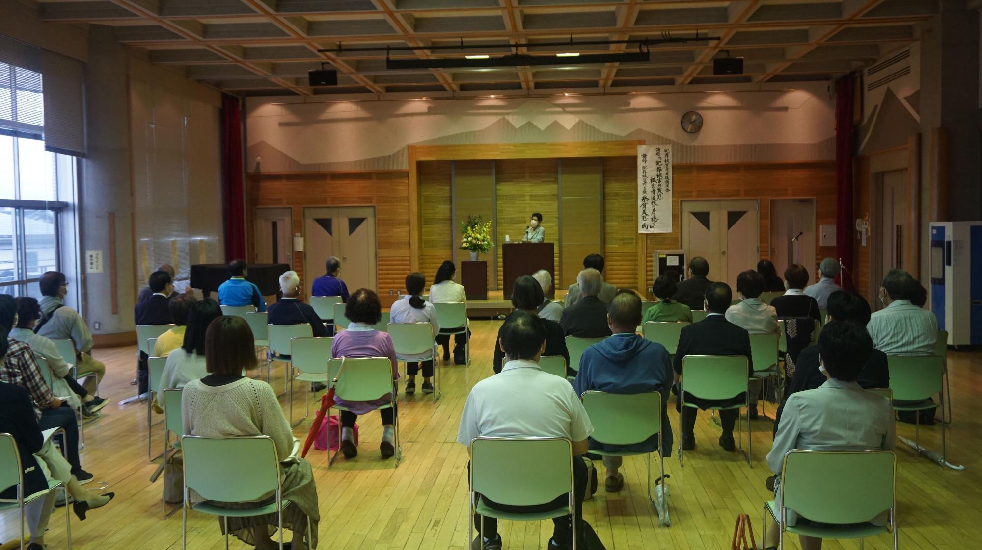木造の格子天井と木製壁面を持つ和風ホールで、演台に立つ講演者と緑色の椅子に着席した多数の聴衆が社会的距離を保って配置されている講演会の写真