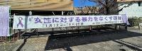 木造施設の外壁に掲げられた「女性に対する暴力をなくす運動11月12日~11月25日」と紫リボンマークを配した白地の長尺横断幕が青空と裸木を背景に設置された啓発広報の写真