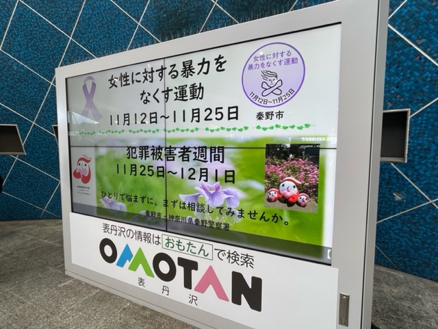 青いタイル壁の前に設置された秦野市の掲示板で、女性に対する暴力をなくす運動と犯罪被害者週間の告知ポスターが重ねて掲示され、下部にOOMOTANのロゴが配置された写真