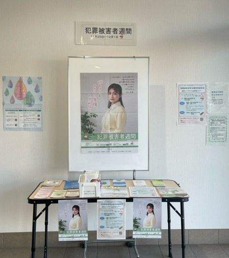 白い壁面に「犯罪被害者週間」の表示と女性が写った啓発ポスターが掲示され、その下の折りたたみ机上に複数のパンフレットや資料が整然と並べられた広報展示コーナーの写真