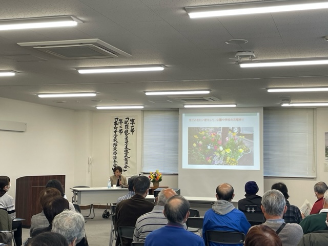 白い壁と蛍光灯照明の会議室で、講師が演台とスクリーンを使い花の写真を投影しながら講演し、多数の参加者が椅子に座って聞き入る講演会の写真