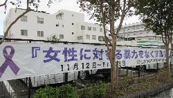 白い建物と街路樹を背景に公共施設のフェンスに掲げられた「女性に対する暴力をなくす運動 11月12日~11月25日」と紫のリボンマークを大きく表示した女性支援啓発の横断幕写真
