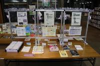 図書館の木製テーブルに女性支援関連書籍・パンフレット・ポスターが配架され、背後の白いパーティションに紫リボンと銀色モールで装飾された女性に対する暴力根絶啓発の特設展示コーナー写真