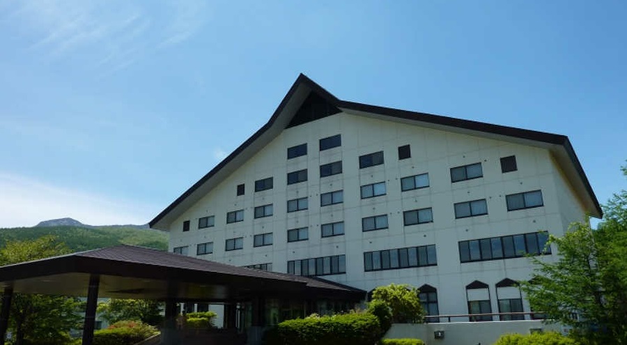 白い外壁と大きな切妻屋根が特徴的なホテルとみられる6階建ての建物を、青空の下、建物の前にある植え込みや右側にある木々を通して下から見上げるように撮影した、遠くに山並みが見える建物の写真