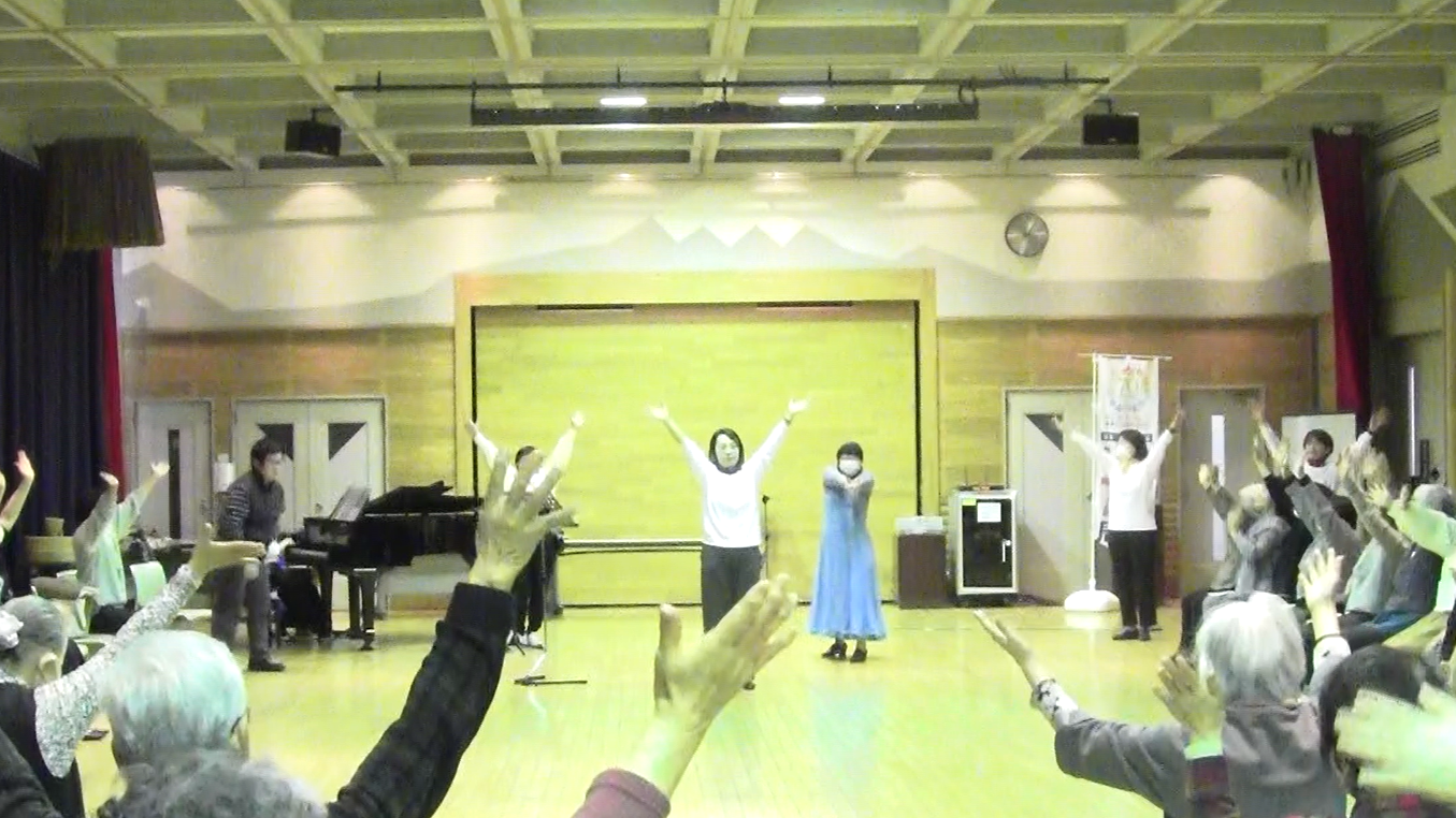 体育館のような広い室内で、中央にいる女性の指導に合わせて多くの参加者が両手を高く上げて体操またはレクリエーション活動をしており、後方にはピアノと白い山並みの背景幕が見える、活動中の集合写真