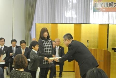 出席者が着席する中、金屏風の前に置かれた演台の前で、スーツ姿の男性が女性と並んで立つ男の子に表彰状を手渡している様子の写真