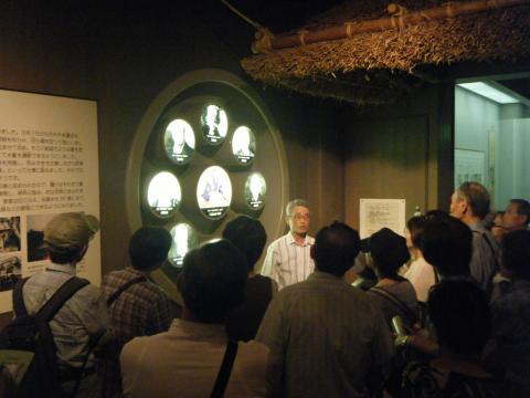 たくさんの参加者が、複数の人物の丸い肖像がディスプレイされた展示物の前に集まり、係の男性の説明を聞いている写真