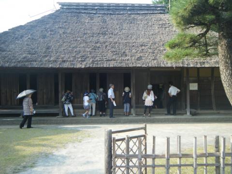 萱葺き屋根の古い日本家屋の前で、たくさんの人々が並んで見学している様子を写した写真