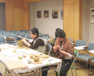 屋内の長机に並んで座った2人の女性が、集中して土器づくりに取り組んでいる様子の写真