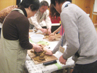 複数の参加者がテーブルを囲んで土器づくりを行っており、エプロンをつけた女性の参加者が男性講師の指導を受けている様子の写真