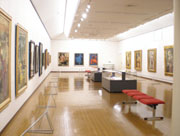 3方の壁にたくさんの絵画作品が展示され、部屋の中央には展示ケースと赤いベンチが置かれた、宮永岳彦美術館の展示室の写真