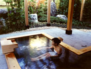 植栽の合間に大きな石が置かれた庭園を臨む、木の枠で囲まれた露天風呂に一人の女性が入浴している、弘法の里湯の写真