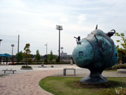 奥に背の高いネットと照明が設置されたグラウンドがあり、手前の石畳の広場にはベンチや植栽が並び、右手前には青みがかった緑色の丸い木のような形のオブジェが置かれているおおね公園の写真