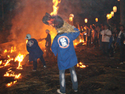 火が燃え盛る地面の前に青い法被を着た数人の男性松明を手にして立ち、その後ろでたくさんの見物客が様子を見守っている、瓜生野百八松明の写真