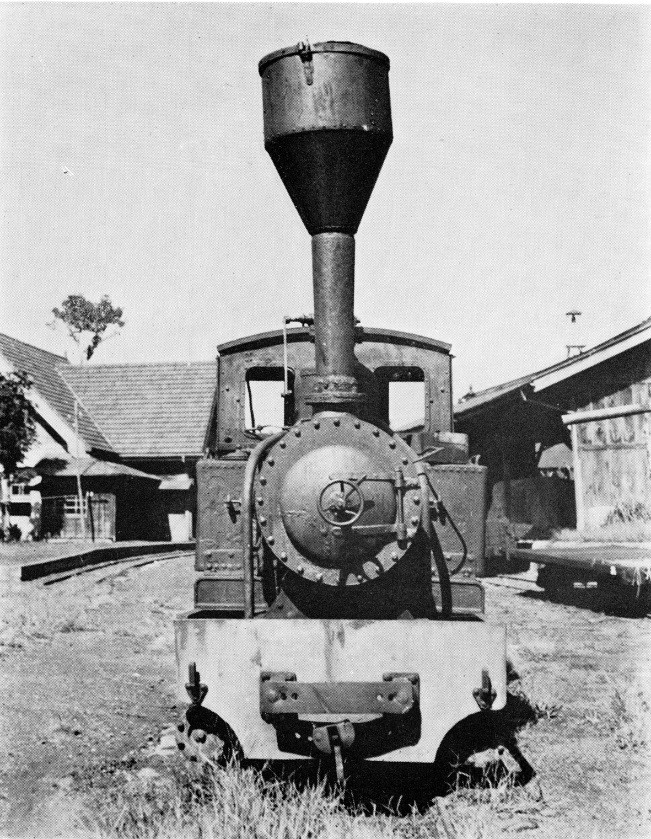 車輪と煙突がついた小型の古い機関車が、木造の建物の前に停まっている軽便鉄道の白黒写真