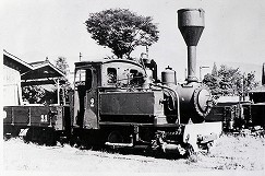 大きな漏斗型の煙突を持つ小型の蒸気機関車とその後ろに貨車が連結されている、線路脇の建物が背景に写り込んだ白黒写真