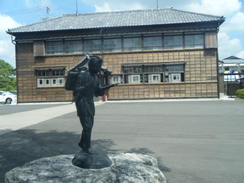 木造建築の建物の手前に、石の台座の上に立つ、薪を背負って本を読みながら歩く姿の二宮尊徳の像が設置されている写真