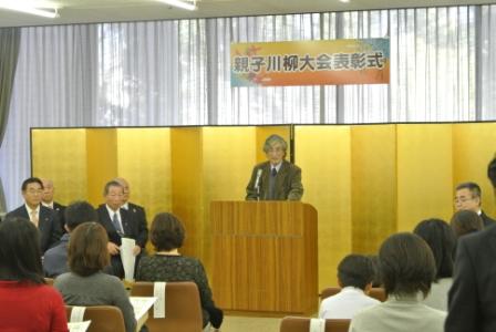 「親子川柳大会表彰式」の横断幕が掲げられた窓の前に設置した金屏風の前で演台に立ったスーツ姿の男性が、着席している出席者たちに話をしている様子の写真